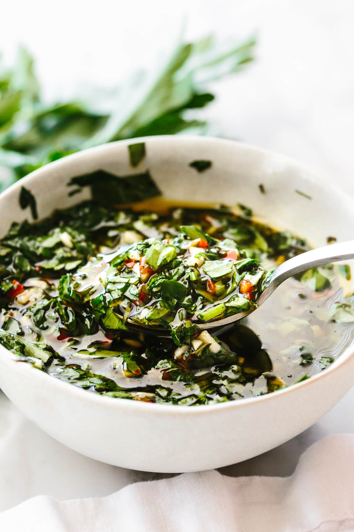 A white bowl of chimichurri sauce next to herbs