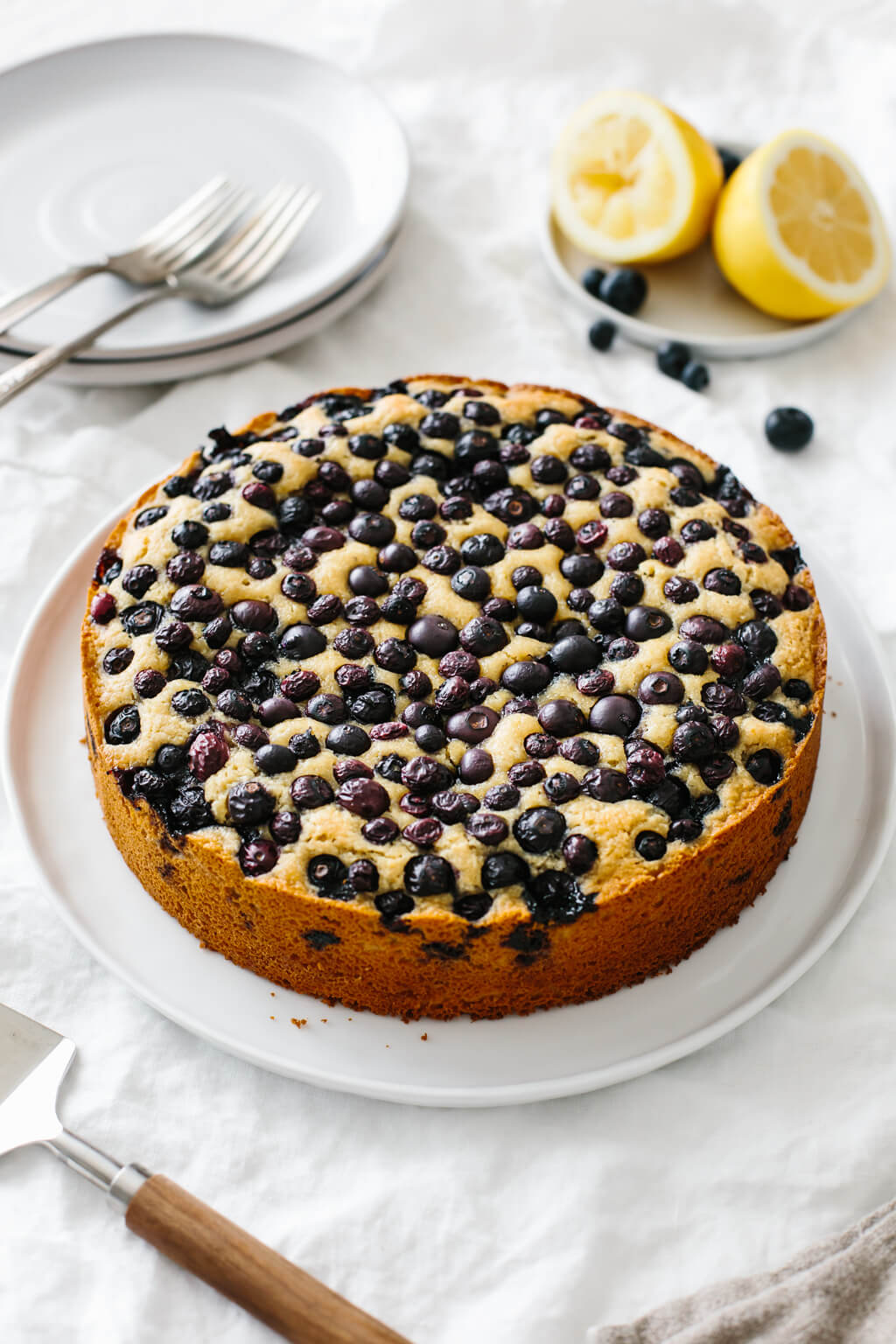 A lemon cake topped with blueberries on a table. A small plate of fresh lemons and blueberries is next to it.